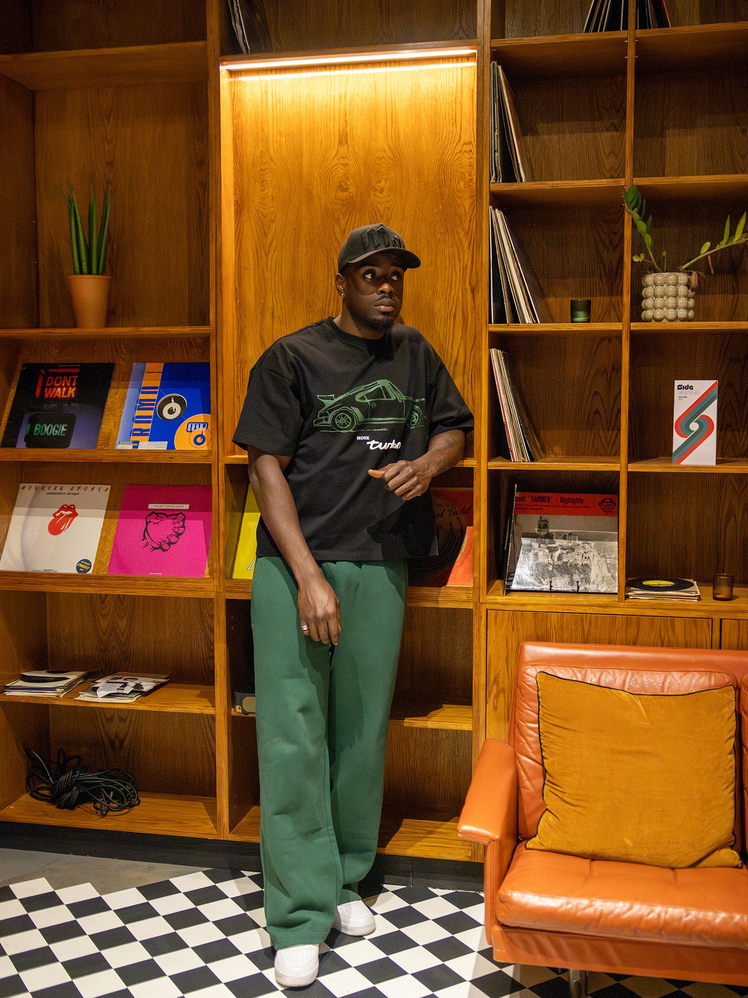 More amoure model wearing a black porsche t-shirt and green amoure pants standing in a room with wooden shelves and a checkered floor.